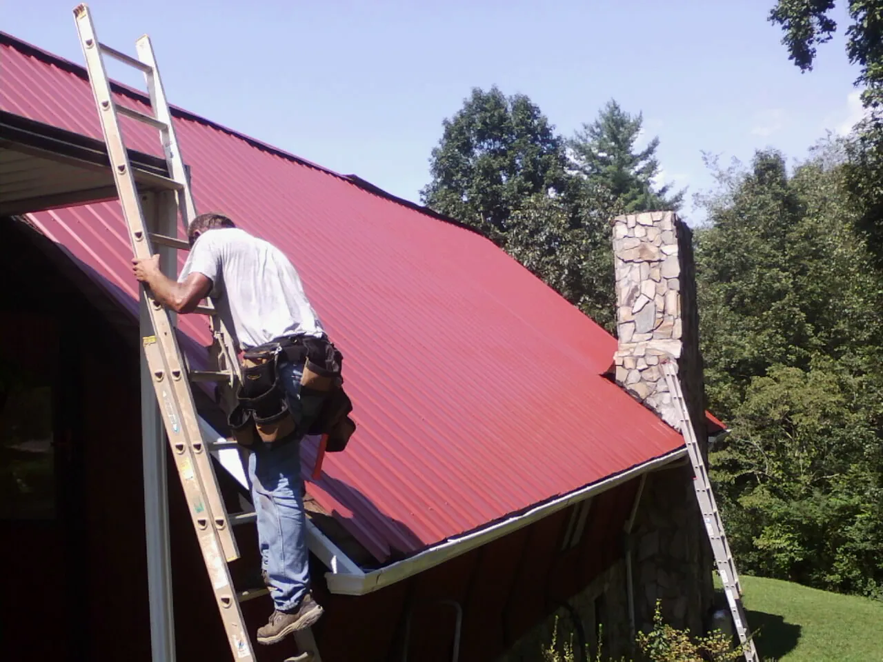 Roofer on a ladder beside a red metal roof and stone chimney — Soffit & Fascia Repair by Roofing Brothers in Curtis